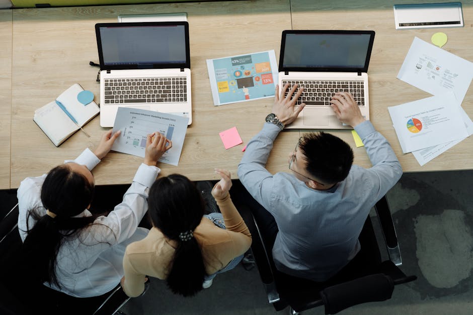 Three colleagues working on laptops and documents at a desk, collaborating on business projects.