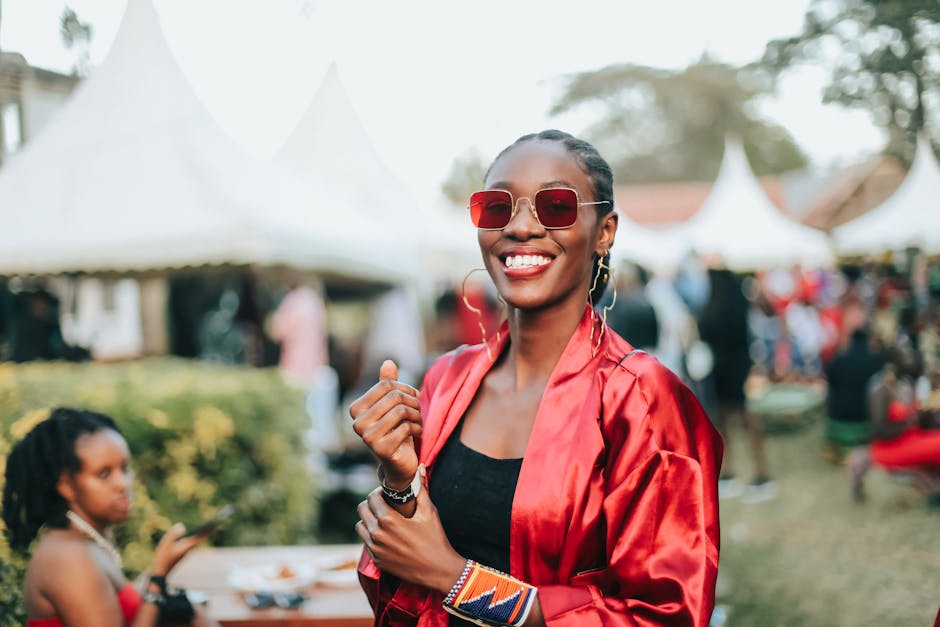 A fashionable woman in a red satin kimono and sunglasses smiles at an outdoor event.